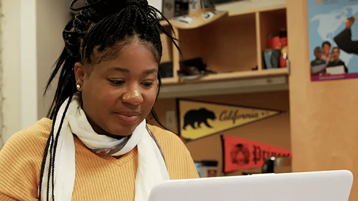 A person wearing a mustard-colored sweater and white scarf works on a laptop in an office-like environment with shelves and posters in the background. A person wearing a mustard-colored sweater and white scarf works on a laptop in an office-like environment with shelves and posters in the background.