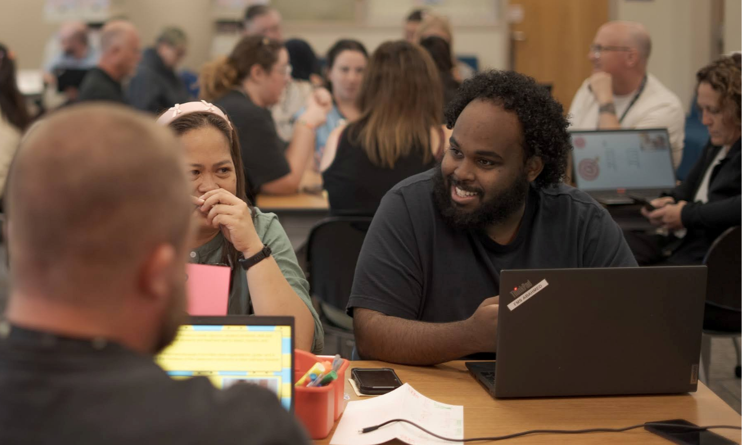 A group of people sitting at tables in a classroom setting, engaged in conversation and working on laptops.