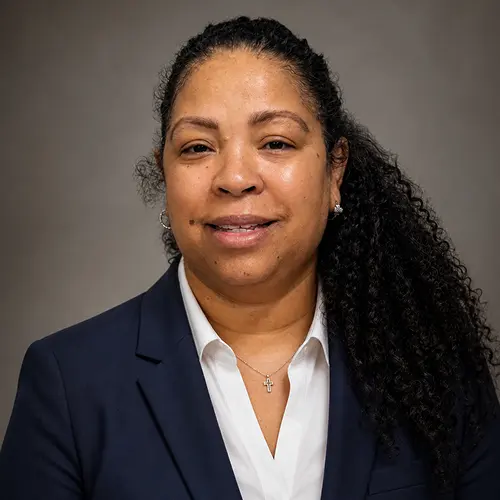 A professional individual wearing a navy blue blazer and white shirt with long curly hair, posing in front of a gray background. A professional individual wearing a navy blue blazer and white shirt with long curly hair, posing in front of a gray background.