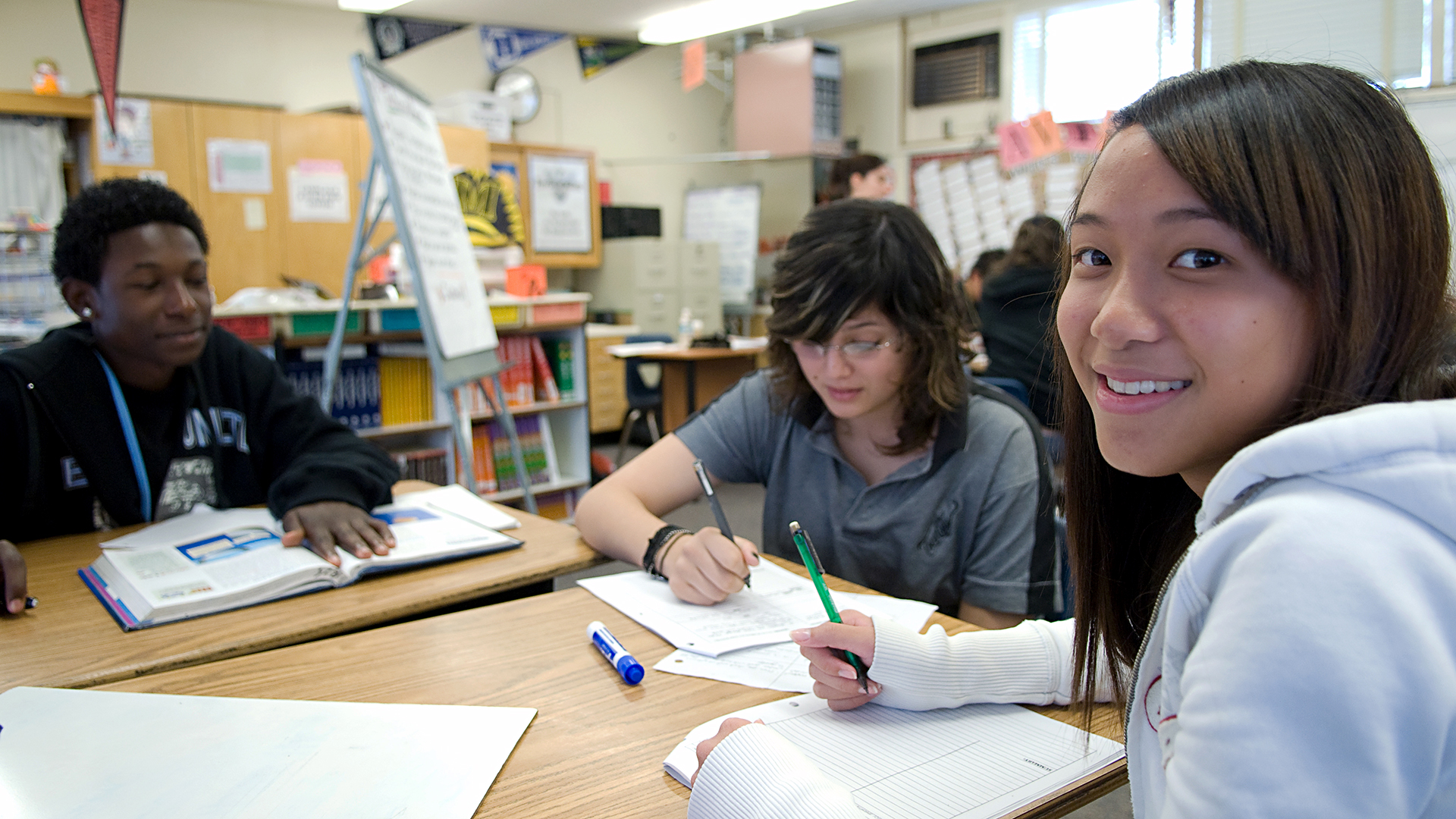 A group of students sitting at desks in a classroom, engaging in study activities with notebooks, textbooks, and pens on the tables. The classroom background includes bookshelves, educational posters, and supplies.