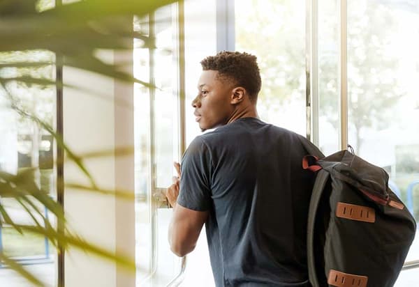 A person wearing a dark t-shirt, carrying a black backpack, standing by a door with glass windows in a brightly lit environment. A person wearing a dark t-shirt, carrying a black backpack, standing by a door with glass windows in a brightly lit environment.