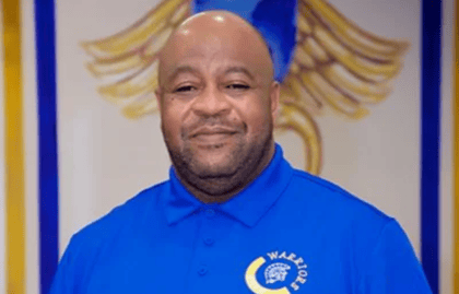 Person in a blue polo shirt with 'Warriors' logo standing in front of a decorative background. Person in a blue polo shirt with 'Warriors' logo standing in front of a decorative background.