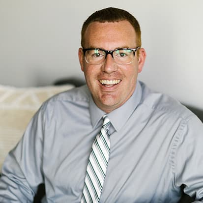 Person wearing a light grey button-up shirt and a striped tie, sitting indoors in a room with light-colored walls. Person wearing a light grey button-up shirt and a striped tie, sitting indoors in a room with light-colored walls.