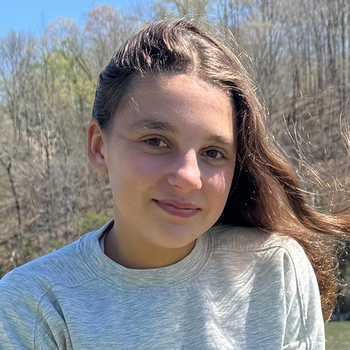 Person with long brown hair wearing a light gray sweater, standing outdoors with trees in the background.