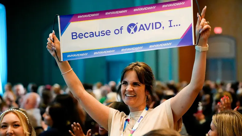 Person holding up a sign with the text 'Because of AVID, I...' in a crowded room. Person holding up a sign with the text 'Because of AVID, I...' in a crowded room.