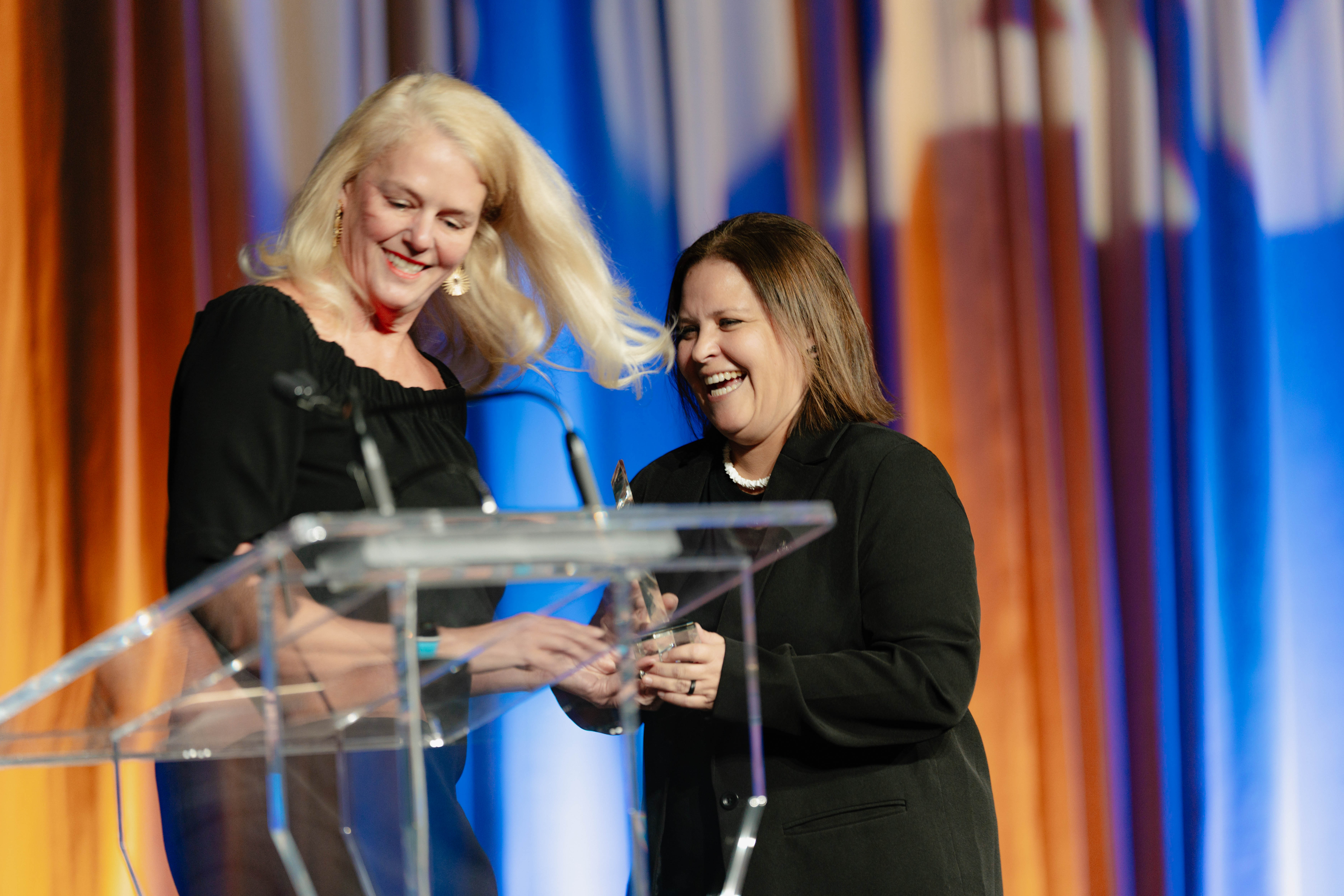 Two individuals standing behind a clear podium on a stage with colorful curtains in the background.
