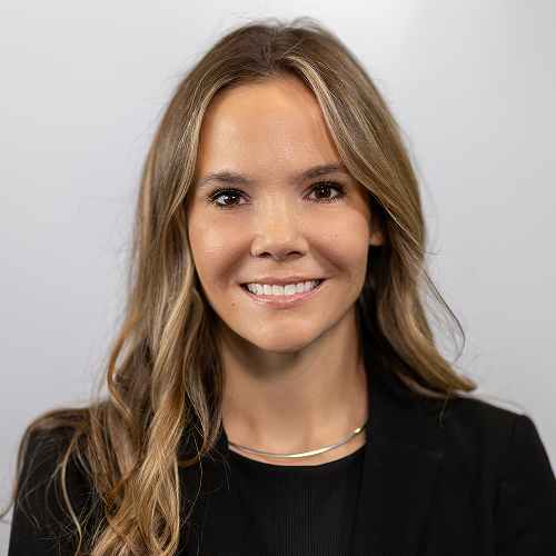 A blurred portrait of a person with long, wavy hair wearing a black blouse and a blazer against a plain background.