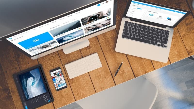 A top view of a wooden desk with a computer monitor, laptop, tablet, smartphone, keyboard, and a pen arranged neatly. A top view of a wooden desk with a computer monitor, laptop, tablet, smartphone, keyboard, and a pen arranged neatly.
