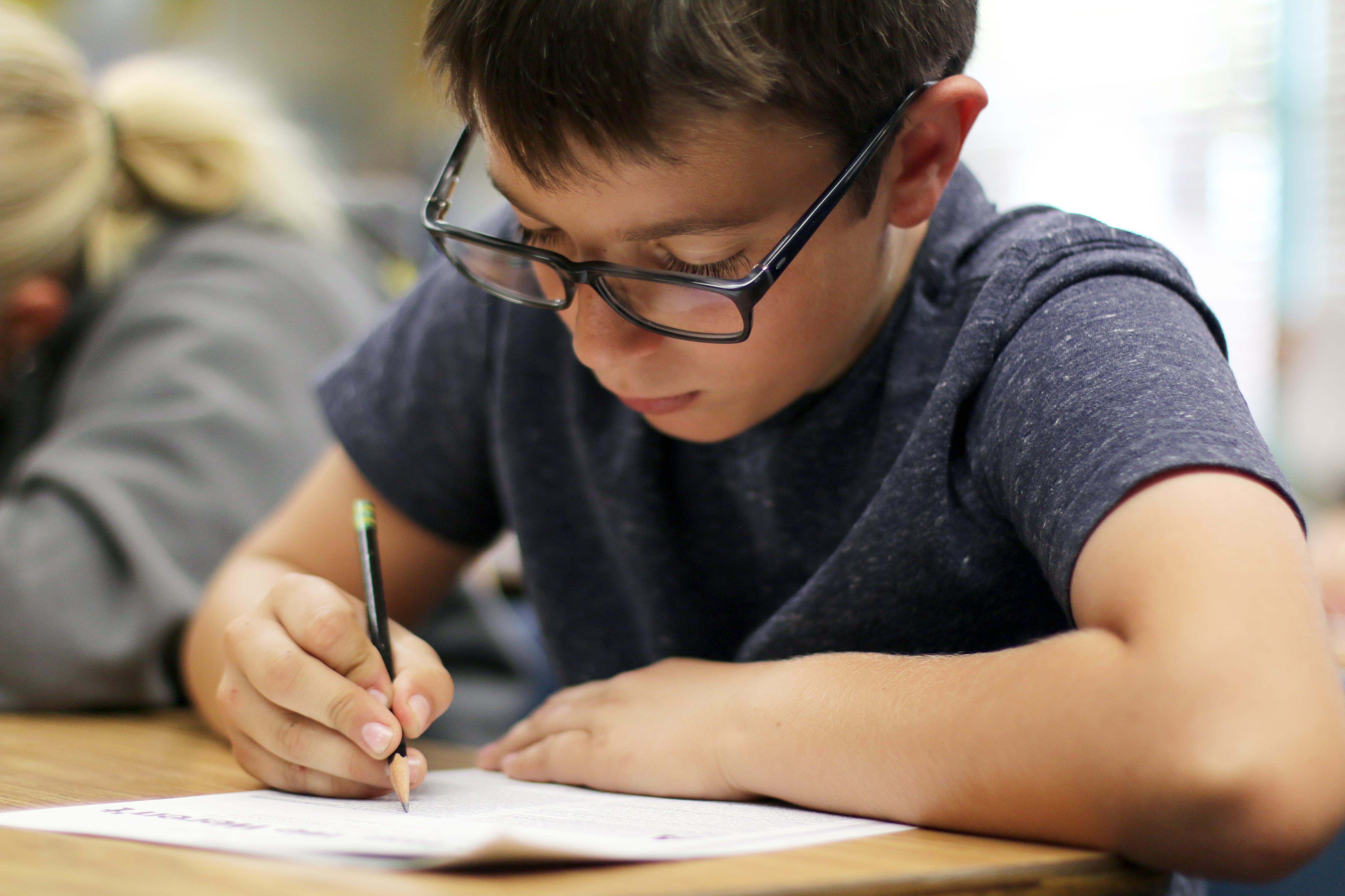 Scholar writing with a pencil on a piece of paper in a classroom setting.