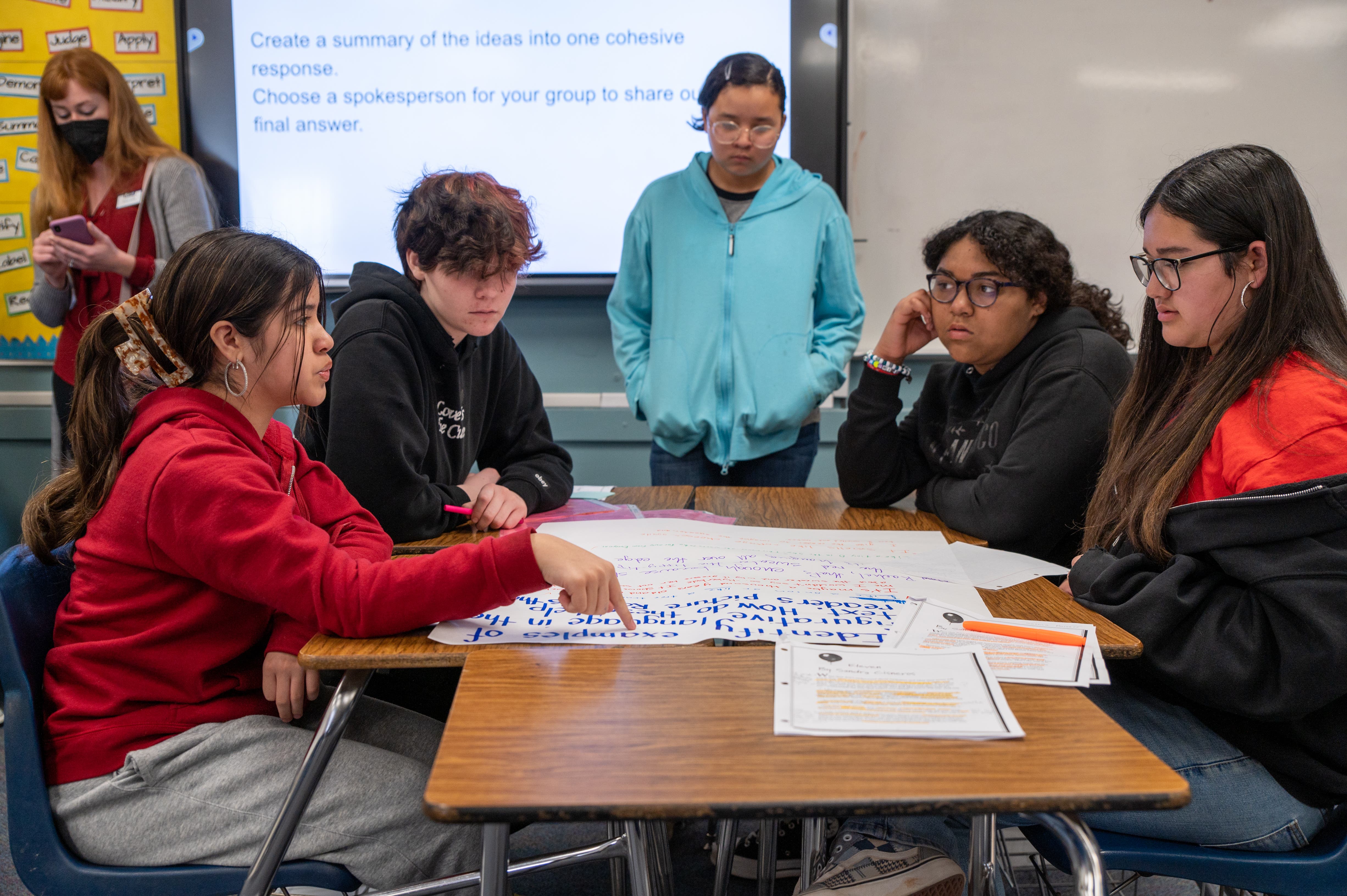Students working together on a project in a classroom setting, with a teacher in the background.