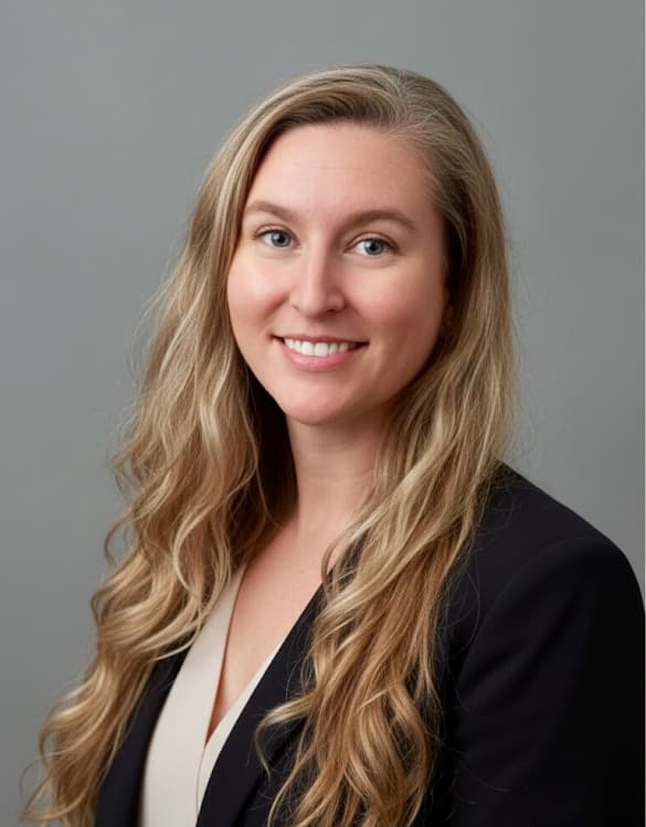 Person with long, wavy hair wearing a dark blazer and light shirt against a gray background. Person with long, wavy hair wearing a dark blazer and light shirt against a gray background.