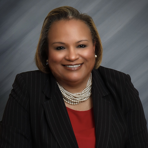 Person wearing a black pinstripe suit jacket, red shirt, pearl necklace, and pearl earrings against a gray background.