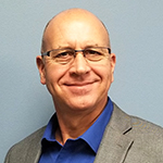 Person wearing a blue shirt and a gray blazer against a plain blue background.