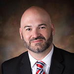 A person dressed in formal attire, including a black suit with a white shirt and a red tie, against a blurred dark background.