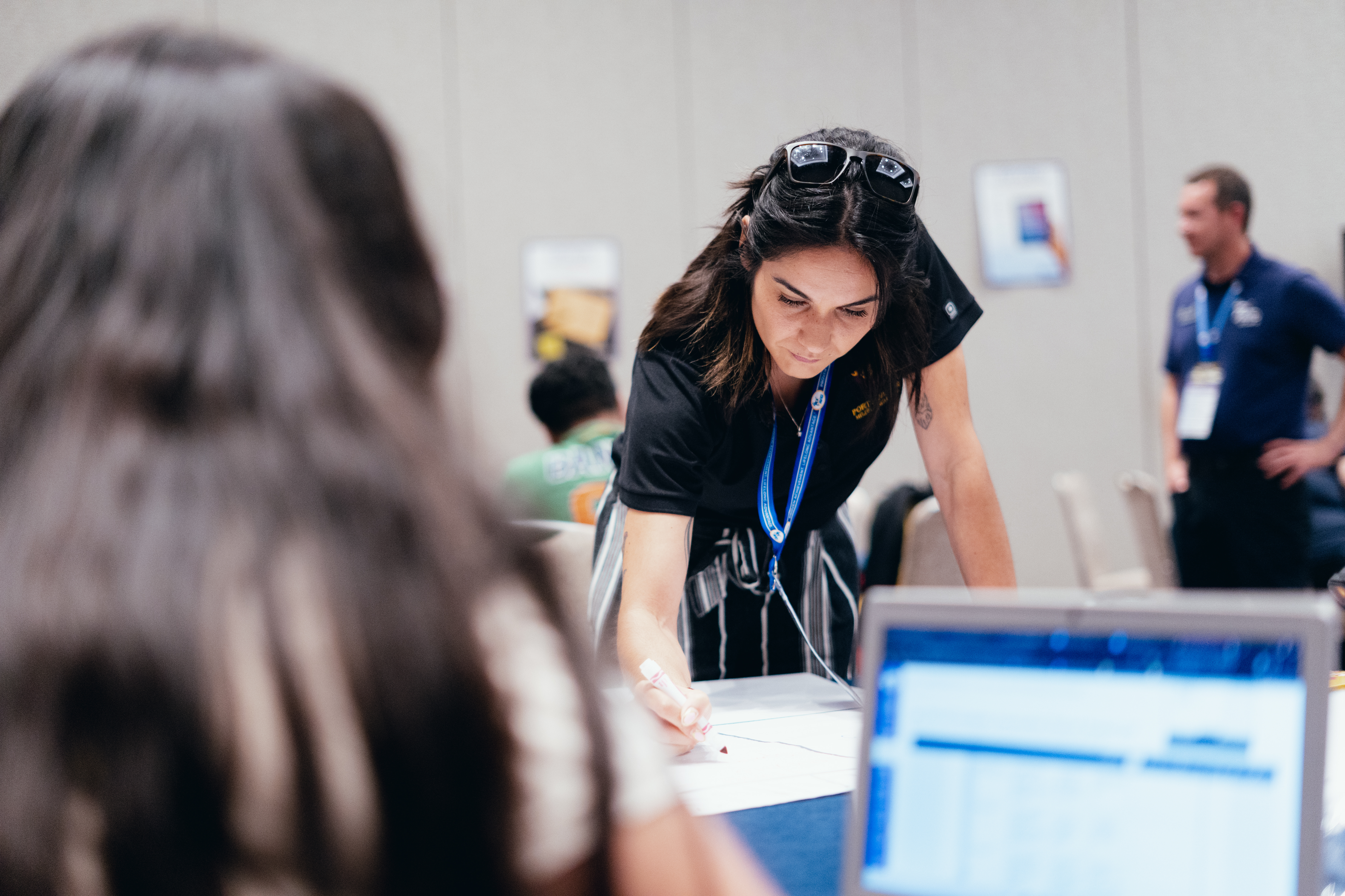 Person with blurred face wearing a black shirt and ID badge leaning over a table with people in the background.