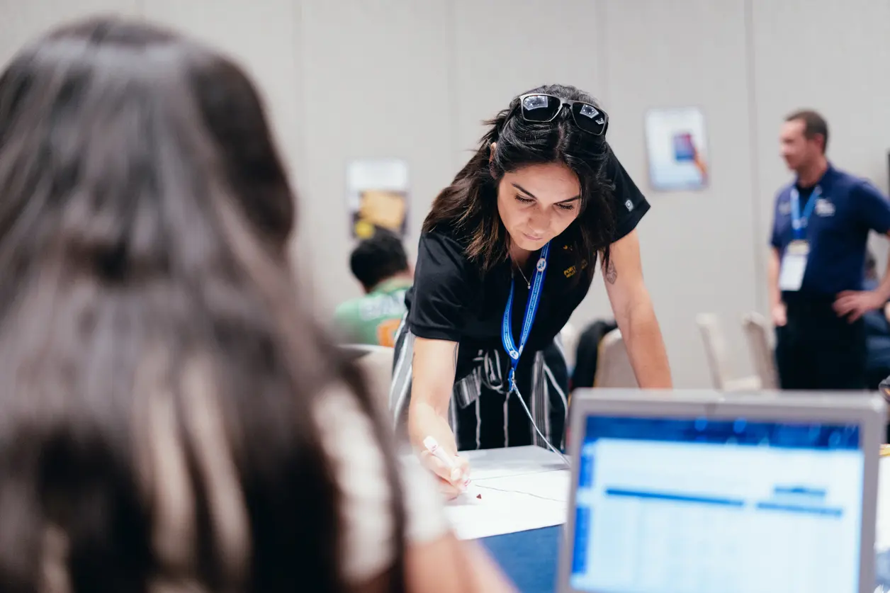 Person with blurred face wearing a black shirt and ID badge leaning over a table with people in the background. Person with blurred face wearing a black shirt and ID badge leaning over a table with people in the background.