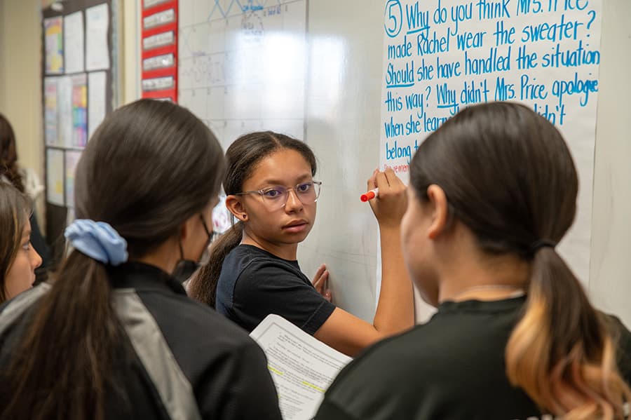 Students working together in a classroom, one writing on a whiteboard and others holding papers. Students working together in a classroom, one writing on a whiteboard and others holding papers.