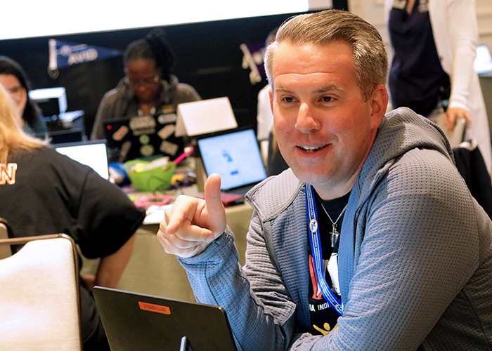 Person in gray hoodie sitting at a table with several laptops and people in the background.