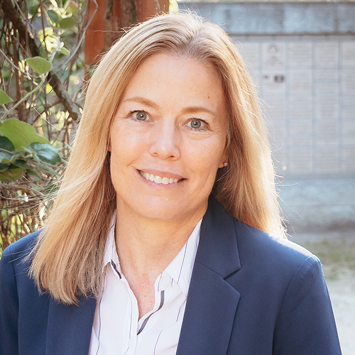 Person with long hair wearing a suit jacket, posing outdoors with greenery in the background.