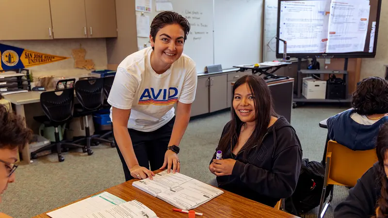 An instructor in a t-shirt assists a student holding a glue stick in a classroom with a desk, chairs, and educational materials in the background. An instructor in a t-shirt assists a student holding a glue stick in a classroom with a desk, chairs, and educational materials in the background.
