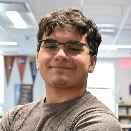 Person standing indoors with blurred face, wearing a patterned long-sleeve shirt. Office or classroom background.
