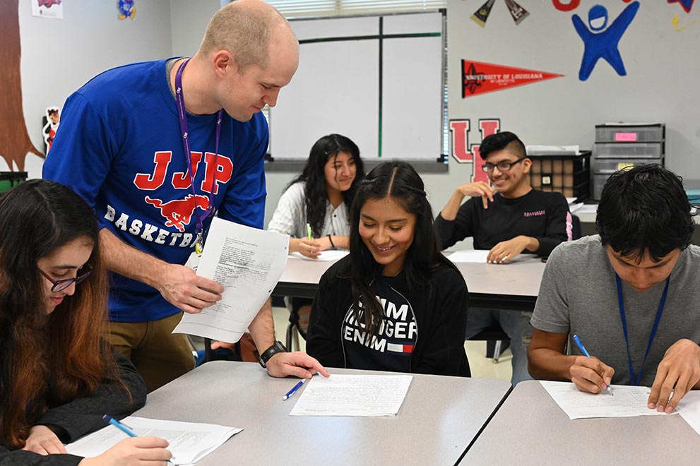 A person in a blue shirt assists a student while others work at their desks in a classroom.