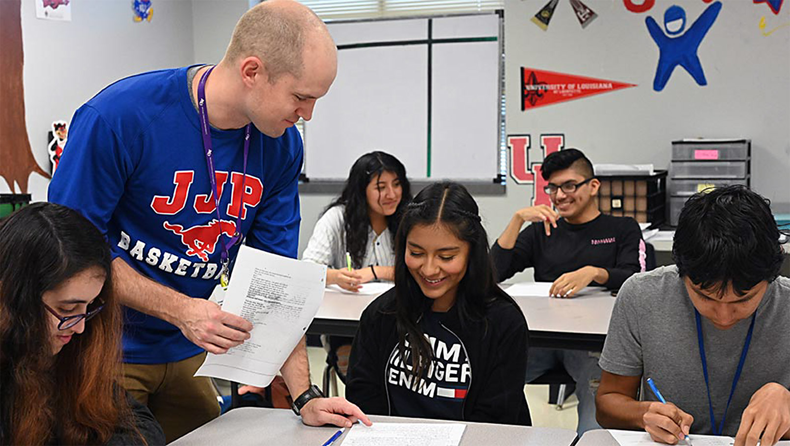 A person in a blue shirt assists a student while others work at their desks in a classroom.