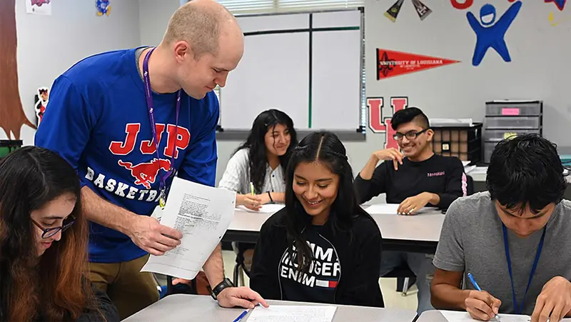 A person in a blue shirt assists a student while others work at their desks in a classroom. A person in a blue shirt assists a student while others work at their desks in a classroom.