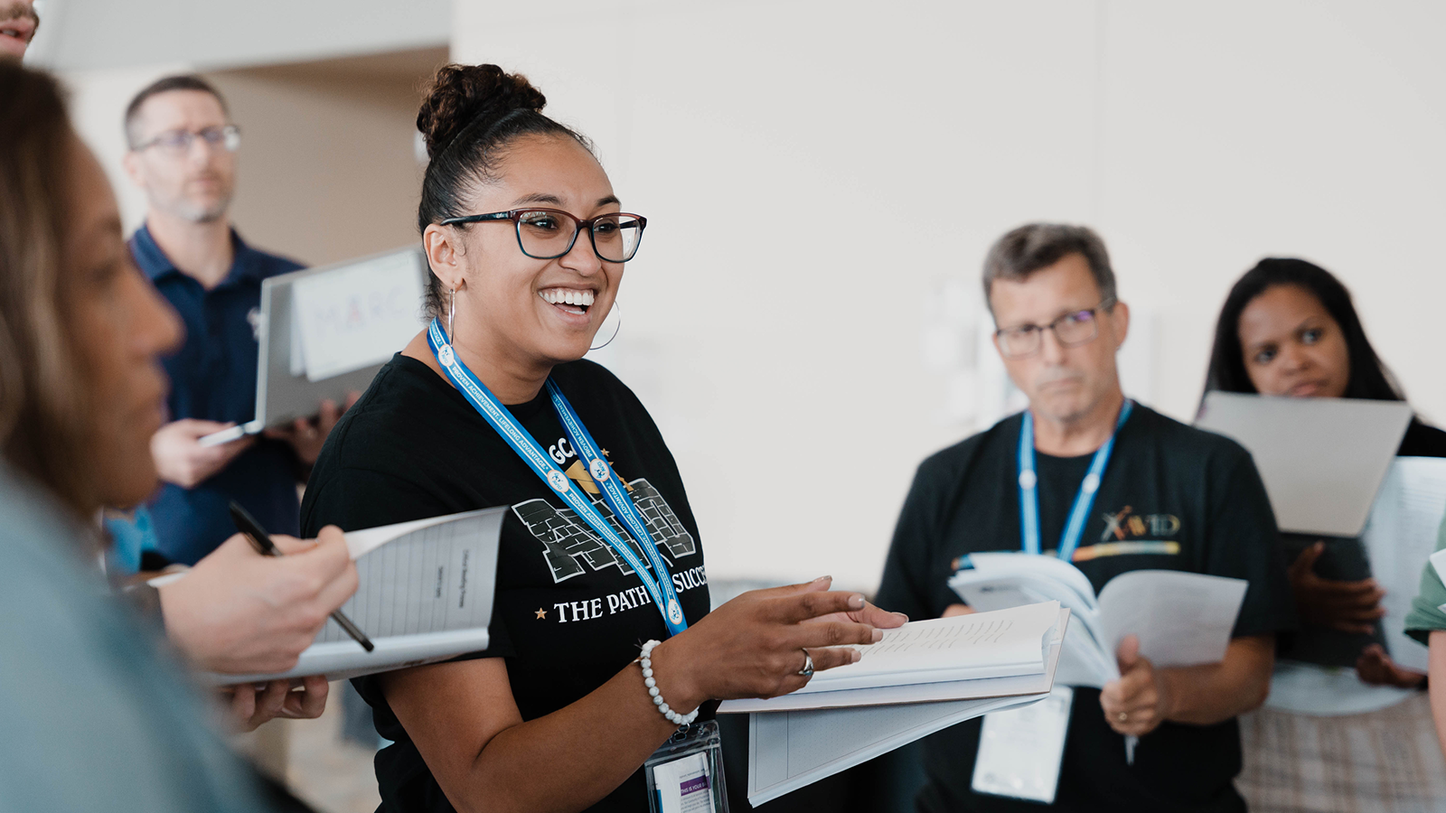 Group of people in a discussion, holding papers and notebooks, at an indoor event.