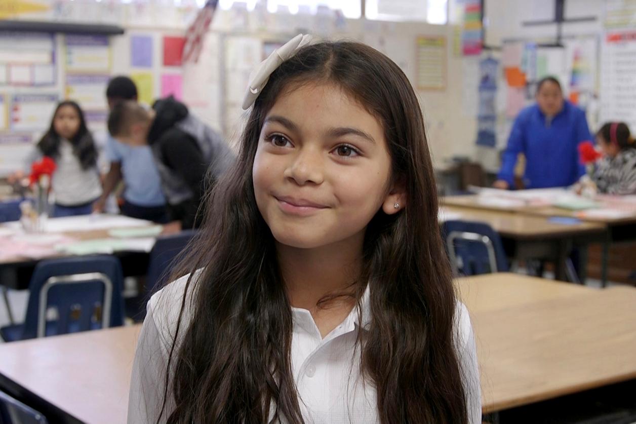 Person with long hair in front of a classroom setting with multiple individuals blurred in the background. Person with long hair in front of a classroom setting with multiple individuals blurred in the background.