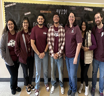 Group photo of seven people standing in front of a blackboard. They are wearing casual outfits, mostly in maroon and white tones.