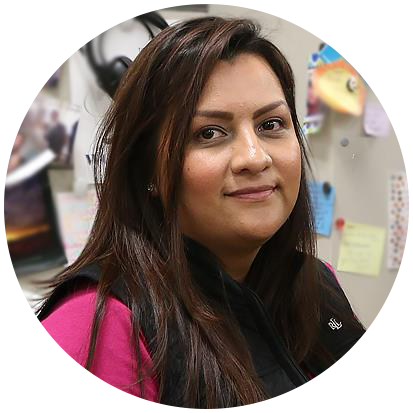 Person with long hair wearing a black vest and pink sleeves in an office setting with note cards and photos on the wall behind them.