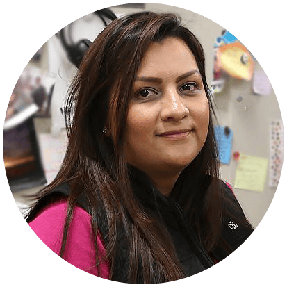 Person with long hair wearing a black vest and pink sleeves in an office setting with note cards and photos on the wall behind them. Person with long hair wearing a black vest and pink sleeves in an office setting with note cards and photos on the wall behind them.