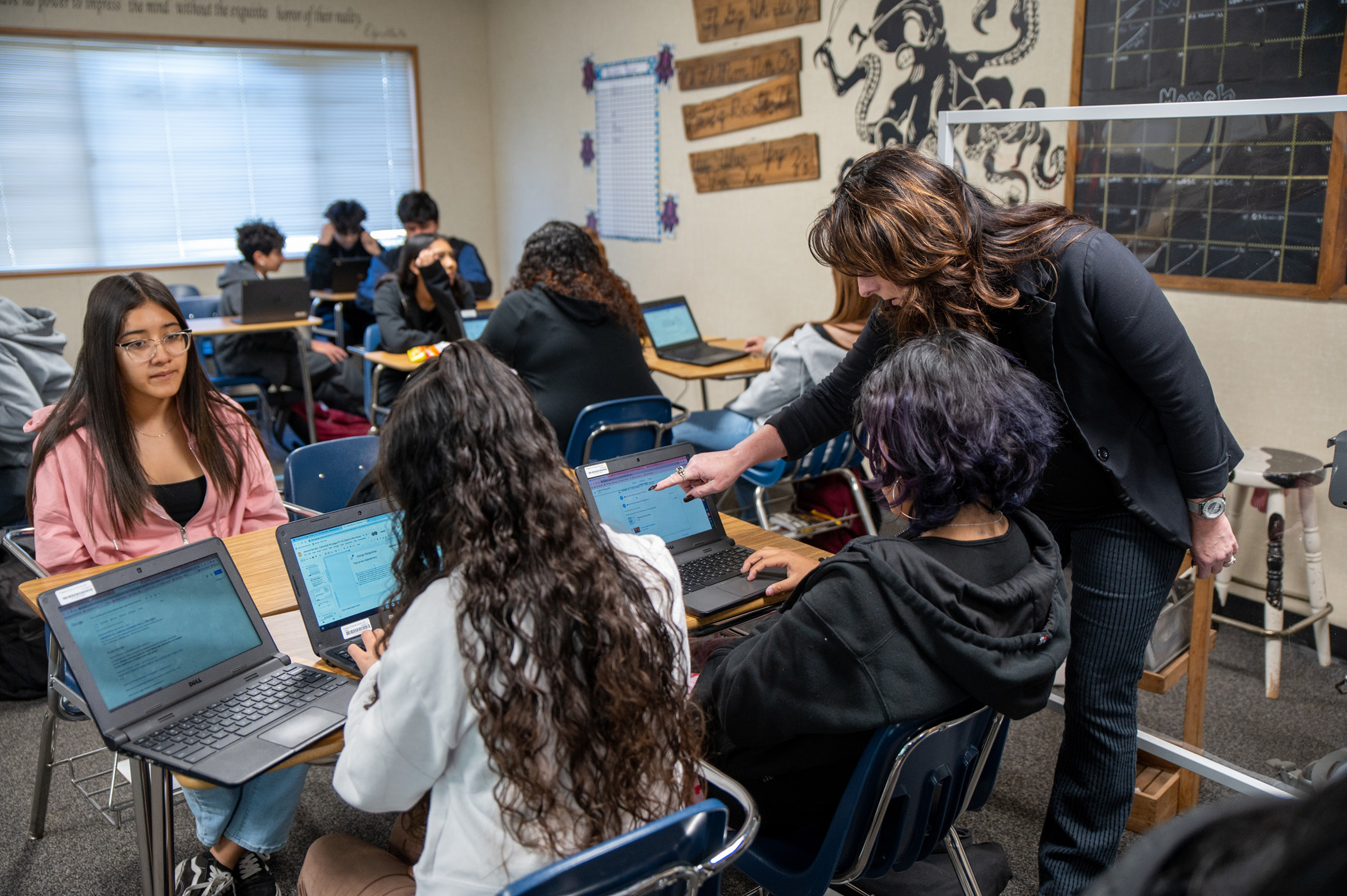 A teacher assisting high school students working on laptops in a classroom.