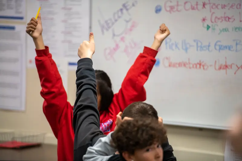 Multiple individuals in a classroom raise their hands, one holding a pencil, in front of a whiteboard with writing. Multiple individuals in a classroom raise their hands, one holding a pencil, in front of a whiteboard with writing.