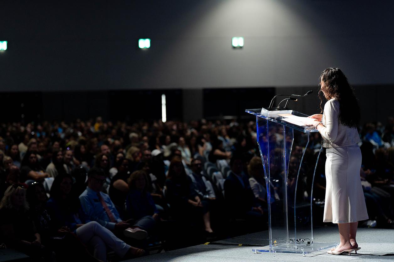 A person in white attire speaking at a transparent podium in front of a large audience A person in white attire speaking at a transparent podium in front of a large audience