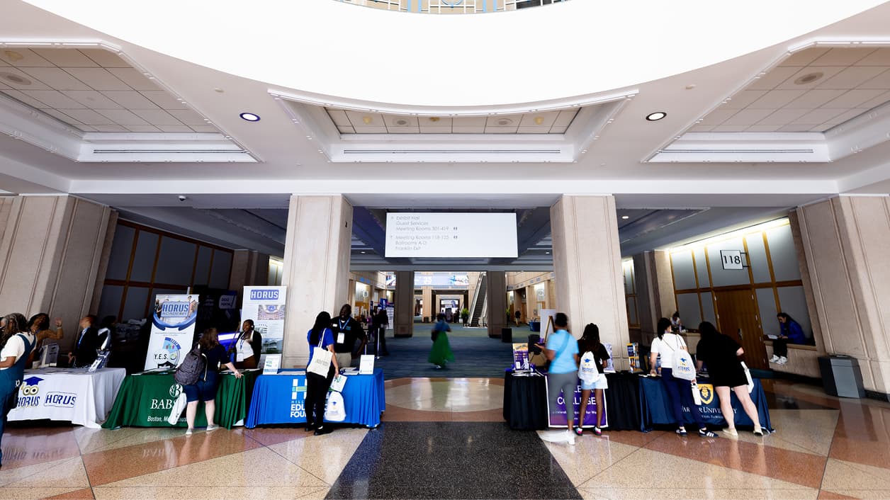 Image of a spacious hallway with booths set up on both sides, featuring various people interacting around the booths. There is a sign overhead that provides directional information for different rooms within the building. Image of a spacious hallway with booths set up on both sides, featuring various people interacting around the booths. There is a sign overhead that provides directional information for different rooms within the building.