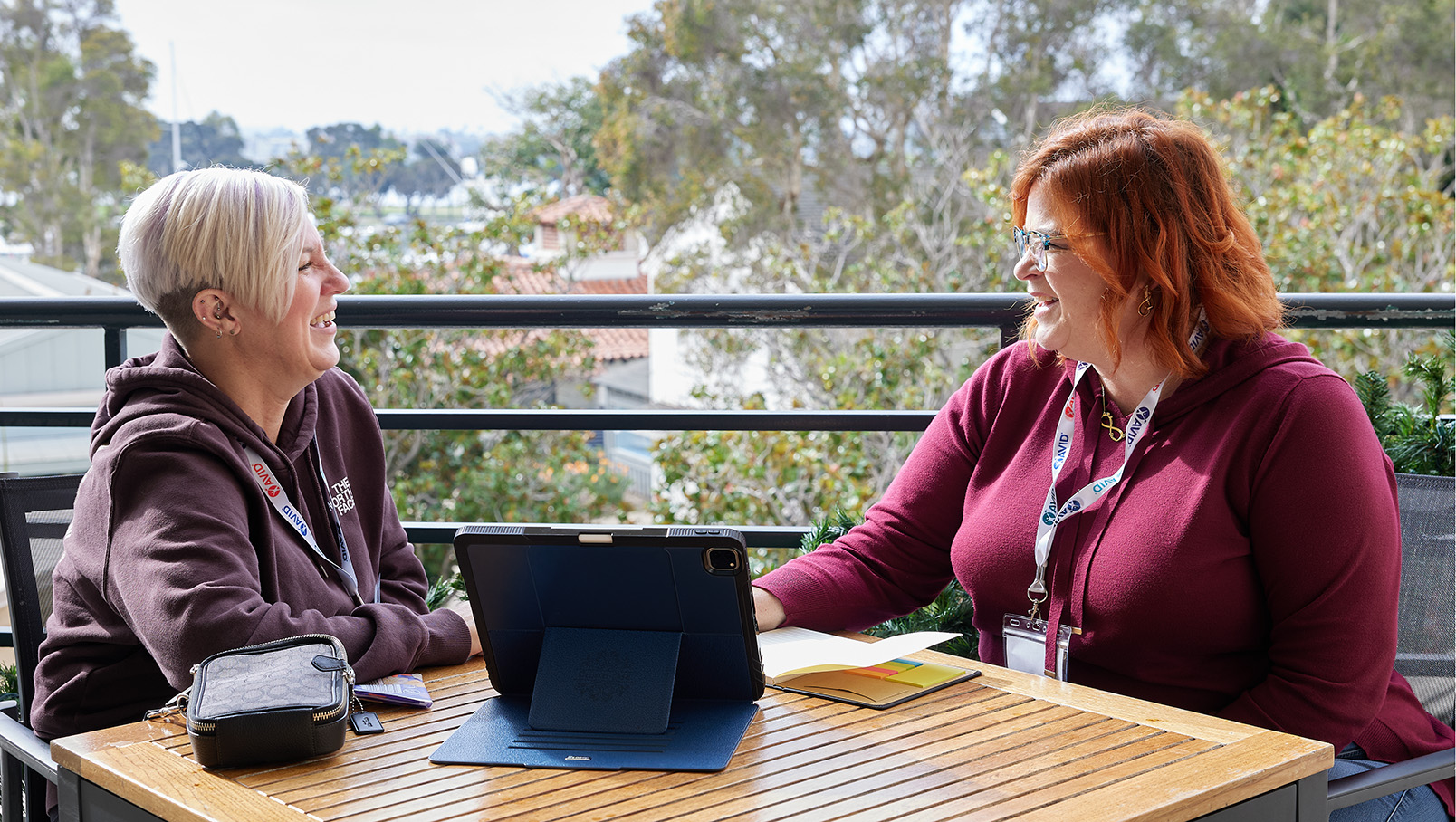 Two people sitting at an outdoor table, smiling and talking. They are wearing lanyards and sweaters, with a tablet and a small bag on the table.