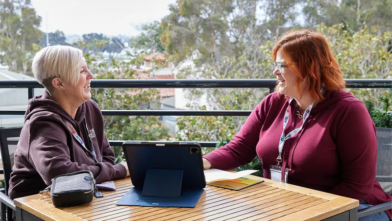 Two people sitting at an outdoor table, smiling and talking. They are wearing lanyards and sweaters, with a tablet and a small bag on the table. Two people sitting at an outdoor table, smiling and talking. They are wearing lanyards and sweaters, with a tablet and a small bag on the table.