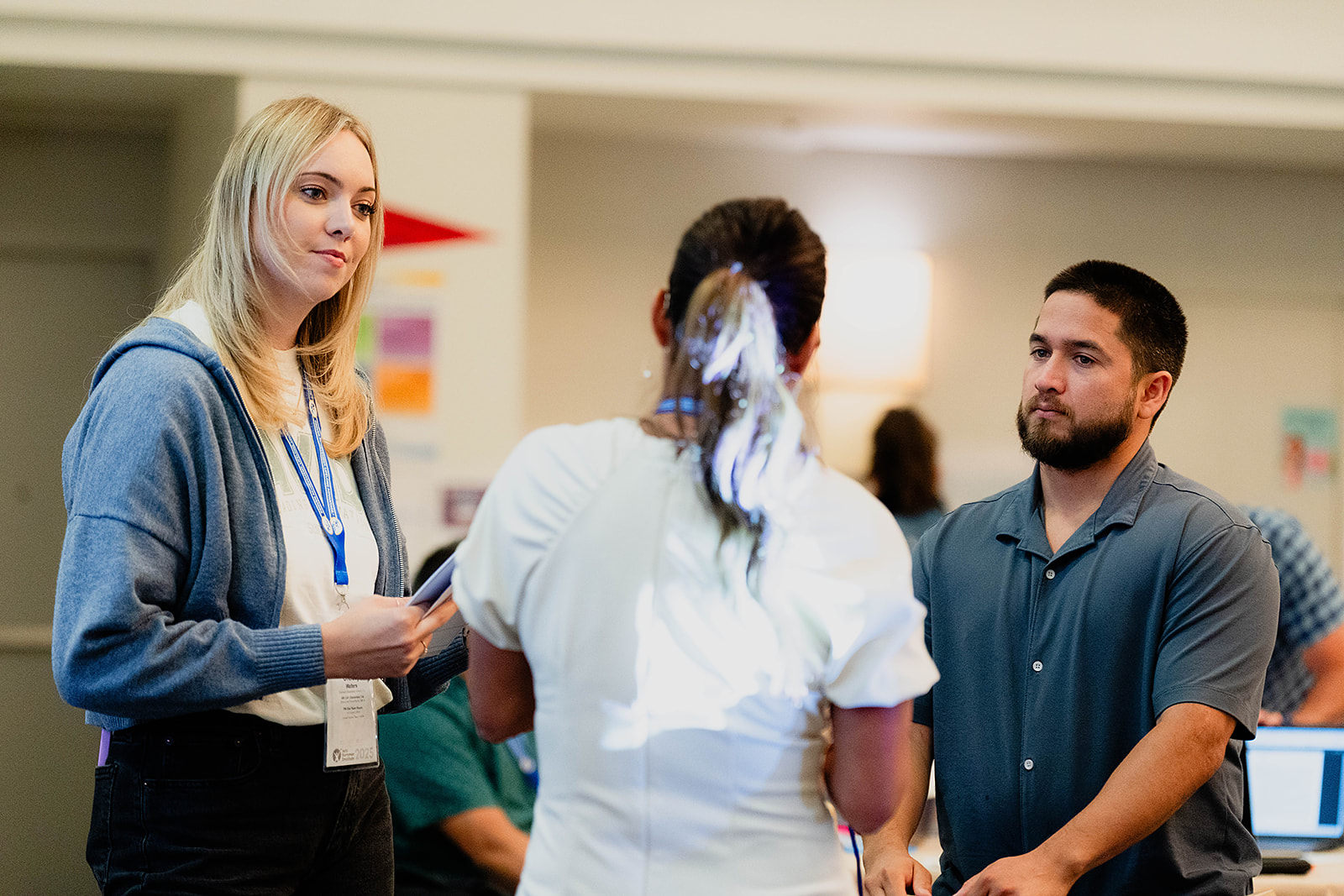 Group of three individuals conversing indoors, one holding a notebook, while the others appear attentive.