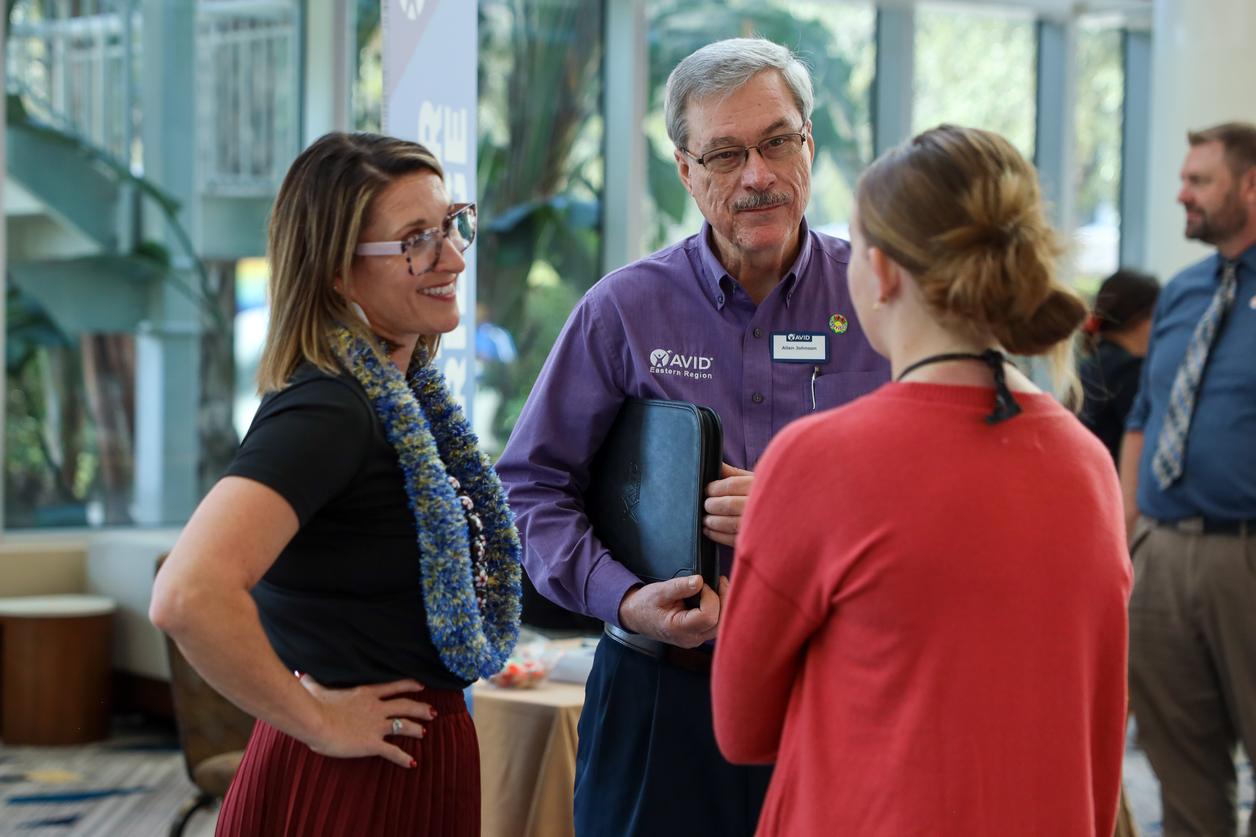A group of people engaged in a conversation at an indoor event, with a man in a purple shirt holding a binder. A group of people engaged in a conversation at an indoor event, with a man in a purple shirt holding a binder.