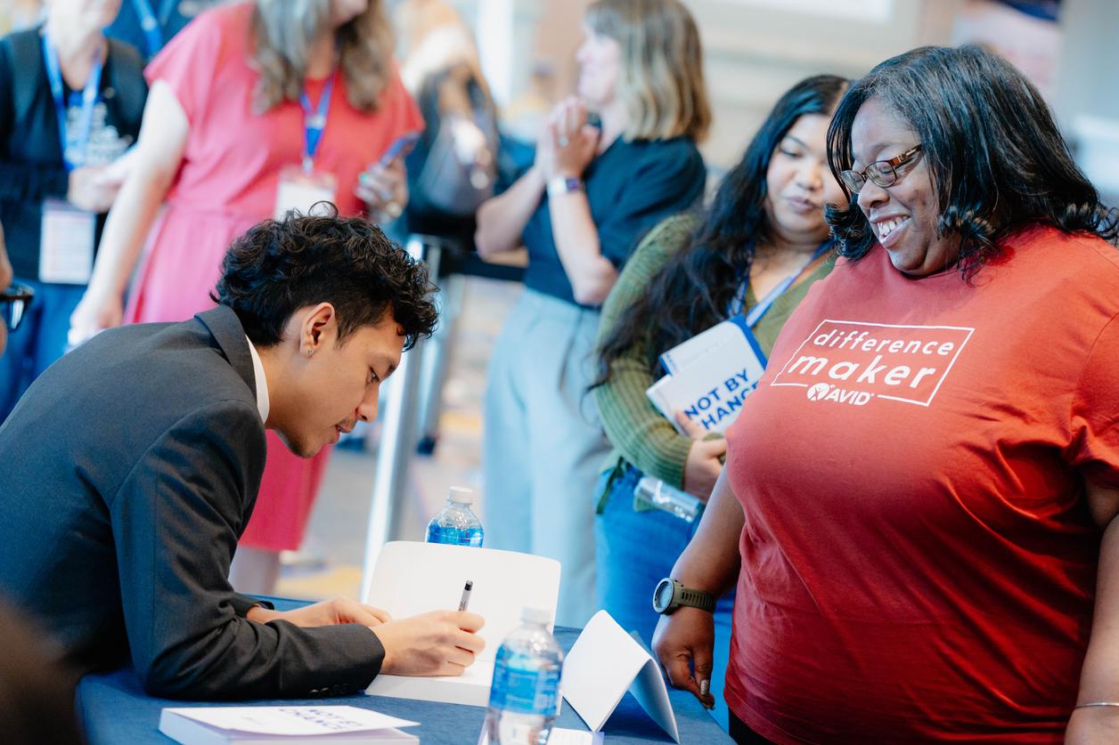 Person sitting at a table and signing something while other people stand nearby. Person sitting at a table and signing something while other people stand nearby.