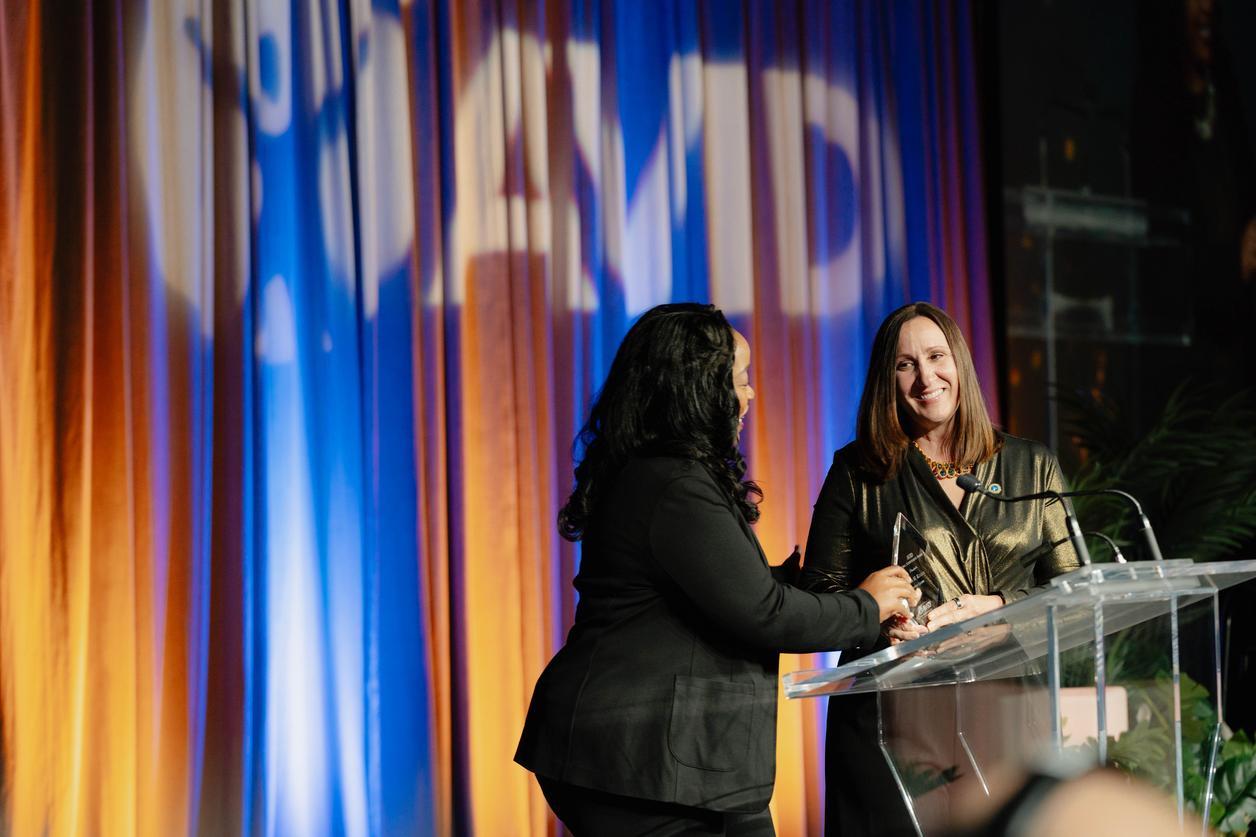 Two individuals at a podium during an event with a backdrop of colorful curtains. Two individuals at a podium during an event with a backdrop of colorful curtains.