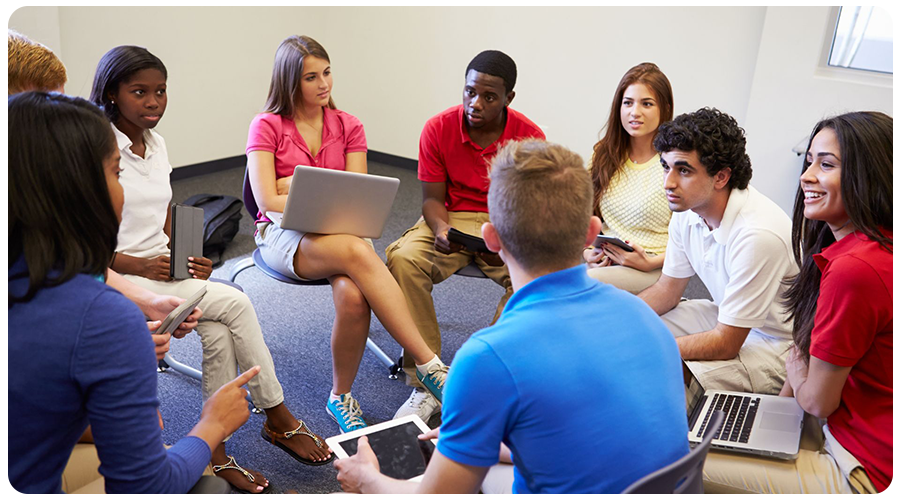 A group of individuals seated in a circular arrangement, engaging in a discussion while using various electronic devices such as laptops and tablets.