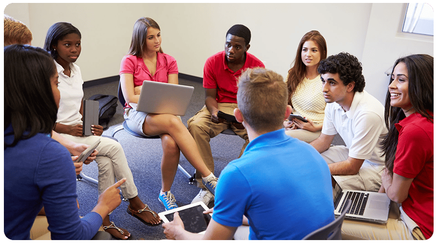 A group of individuals seated in a circular arrangement, engaging in a discussion while using various electronic devices such as laptops and tablets. A group of individuals seated in a circular arrangement, engaging in a discussion while using various electronic devices such as laptops and tablets.