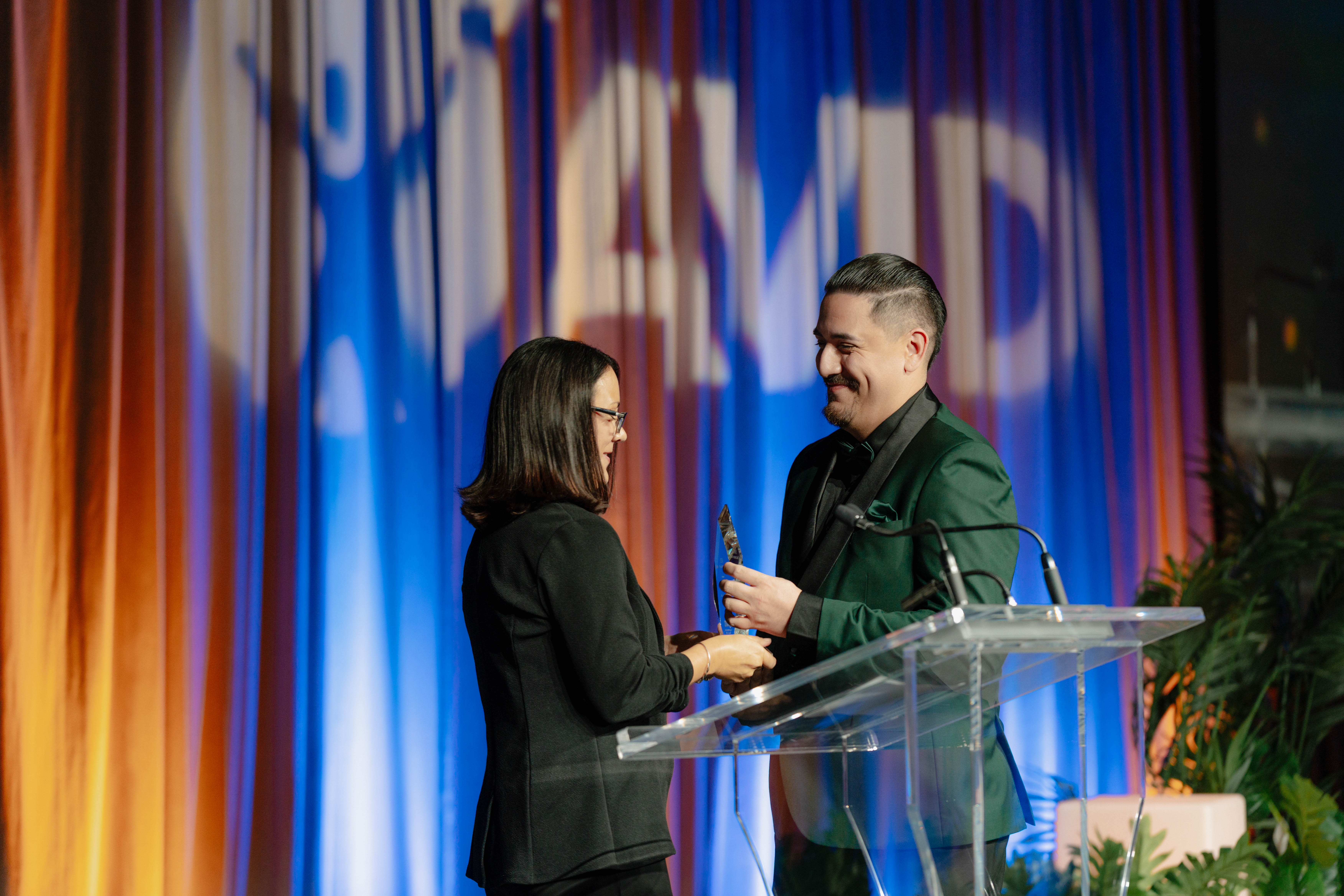 Two individuals stand on stage in front of a podium, with one person holding an award.