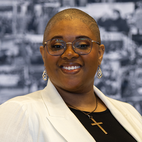 Person wearing a white jacket and black shirt with a cross necklace, background is blurred.