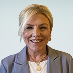 A person with blonde hair wearing a grey blazer and a gold necklace against a light blue background.