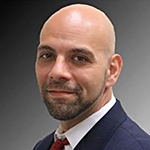 A close-up image of a person wearing a dark suit and a red tie against a gradient gray background.