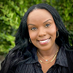 Person with long dark hair wearing a black top and gold necklace with a blurred face standing against a green foliage background.